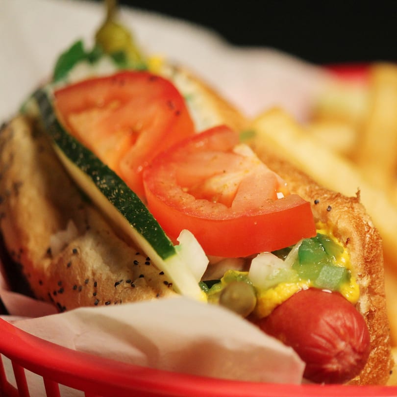 A Chicago-style hot dog with all the fixings, including tomatoes, cucumber, and relish, in a red basket.